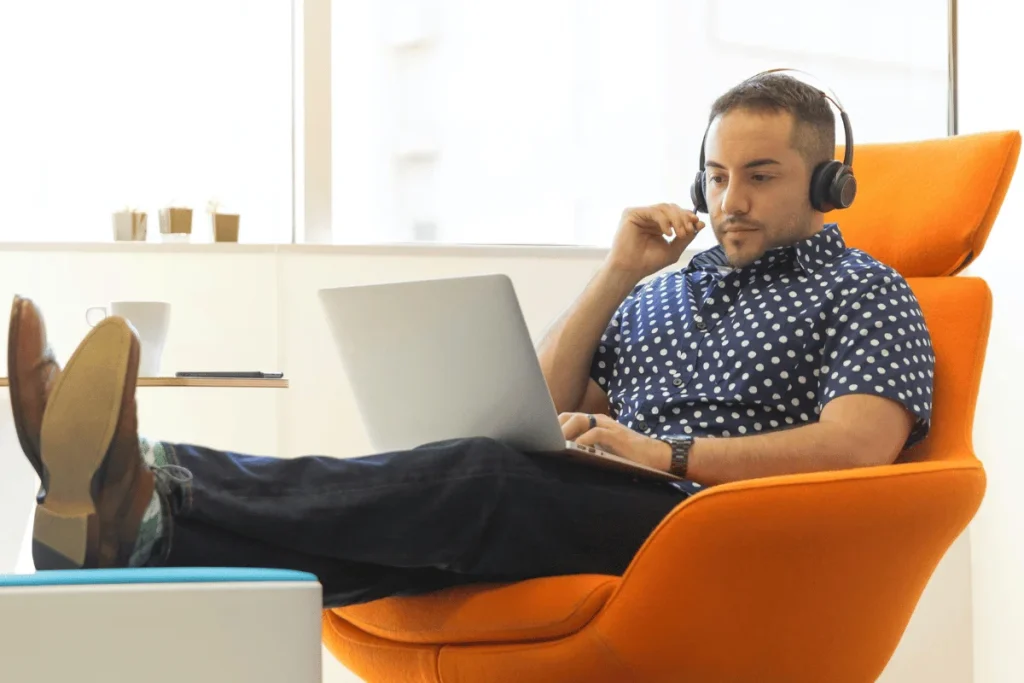 Relaxed man in headphones using laptop while reclining in an orange chair.