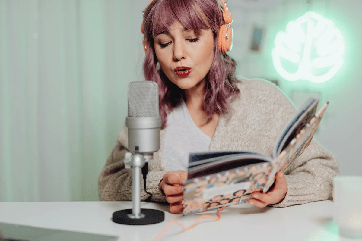 A person with purple hair wears headphones and holds an open magazine while sitting at a table with a microphone. A glowing leaf-shaped light is in the background.