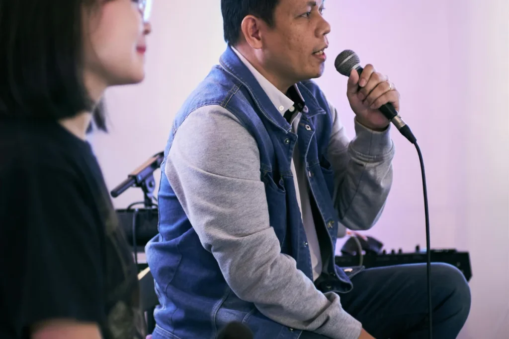 Man holding a microphone, engaged in a discussion, with another person slightly blurred in the foreground.