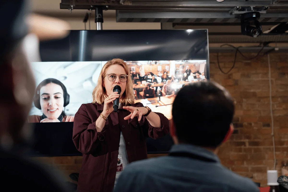 A presenter gestures while speaking into a microphone, with an audience and a large screen displaying a video conference in the background.