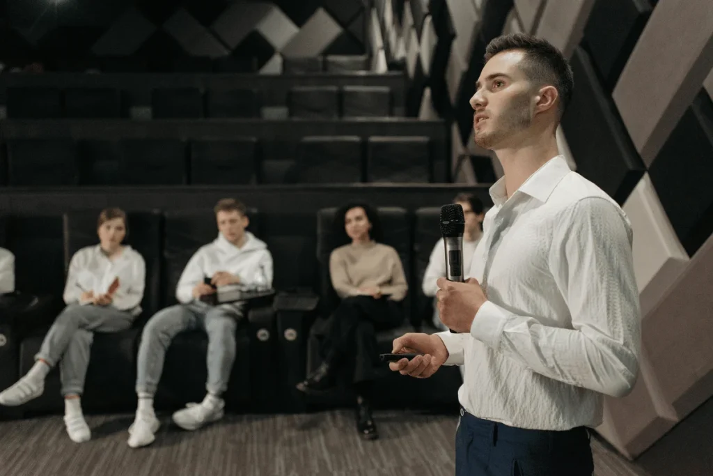 A confident speaker holding a microphone while presenting to an engaged audience in a modern auditorium.