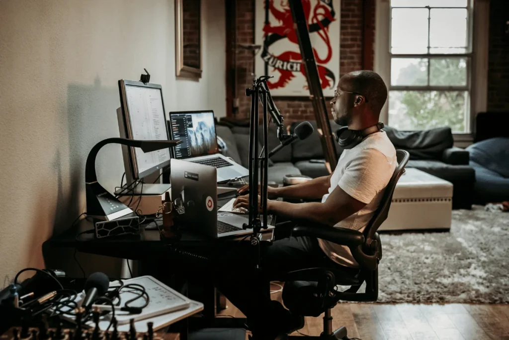 Man seated at a workstation with dual monitors, a podcast microphone, and headphones, recording a podcast.