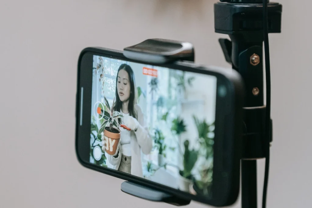 A smartphone mounted on a tripod records a woman presenting a potted plant in a bright indoor setting, suggesting a calm gardening tutorial or livestream.