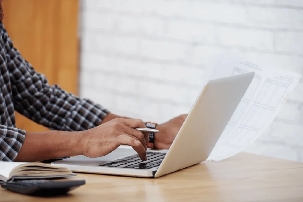 Detail of hands typing on a laptop while reviewing printed spreadsheets.