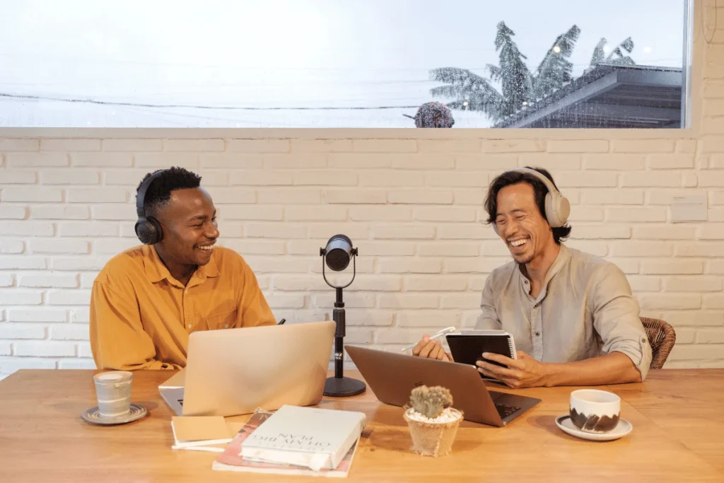 Two men laughing while recording a podcast at a cozy indoor setup.