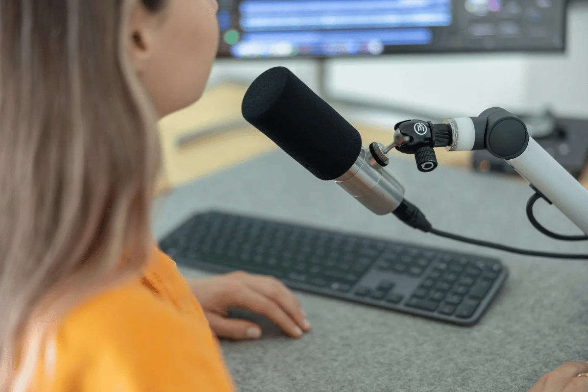 A person speaks into a microphone on an adjustable stand, with a keyboard and computer monitor in the background. The setting suggests a podcast or recording studio.