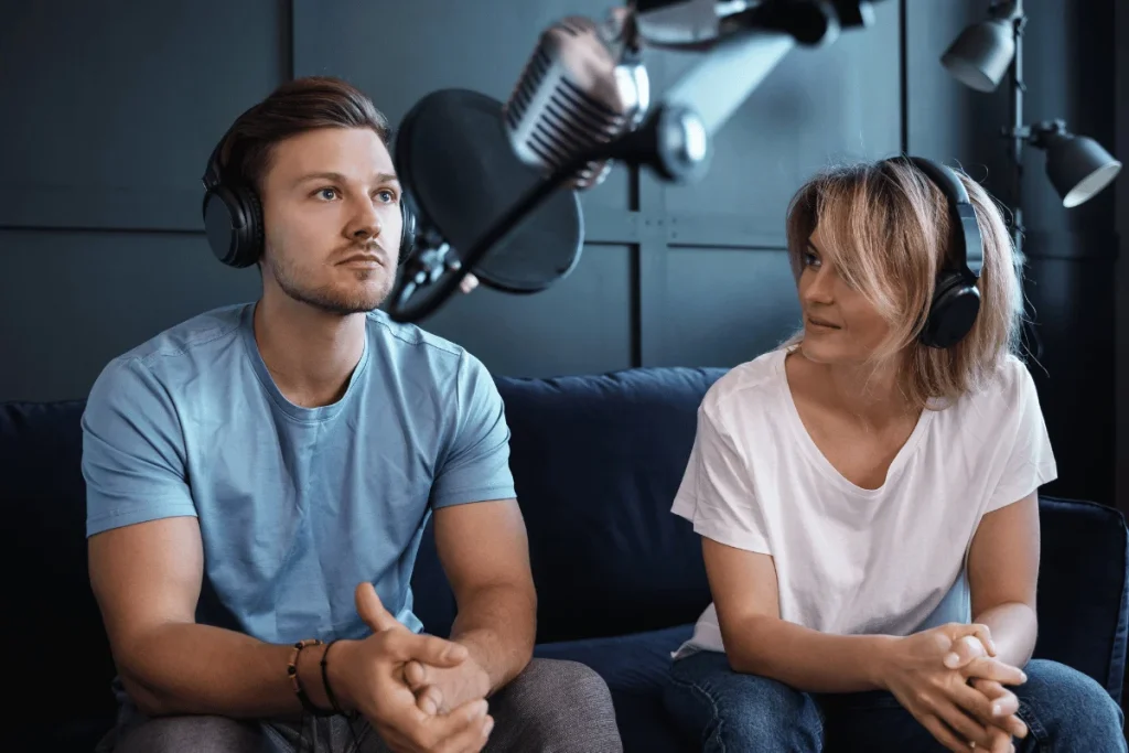 Two podcast hosts wearing headphones sit on a couch, preparing to record with a microphone in a professional studio setup.