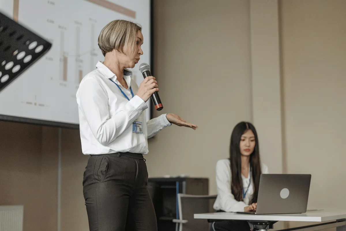 A businesswoman presenting data insights with a microphone, supported by a teammate seated at a desk.