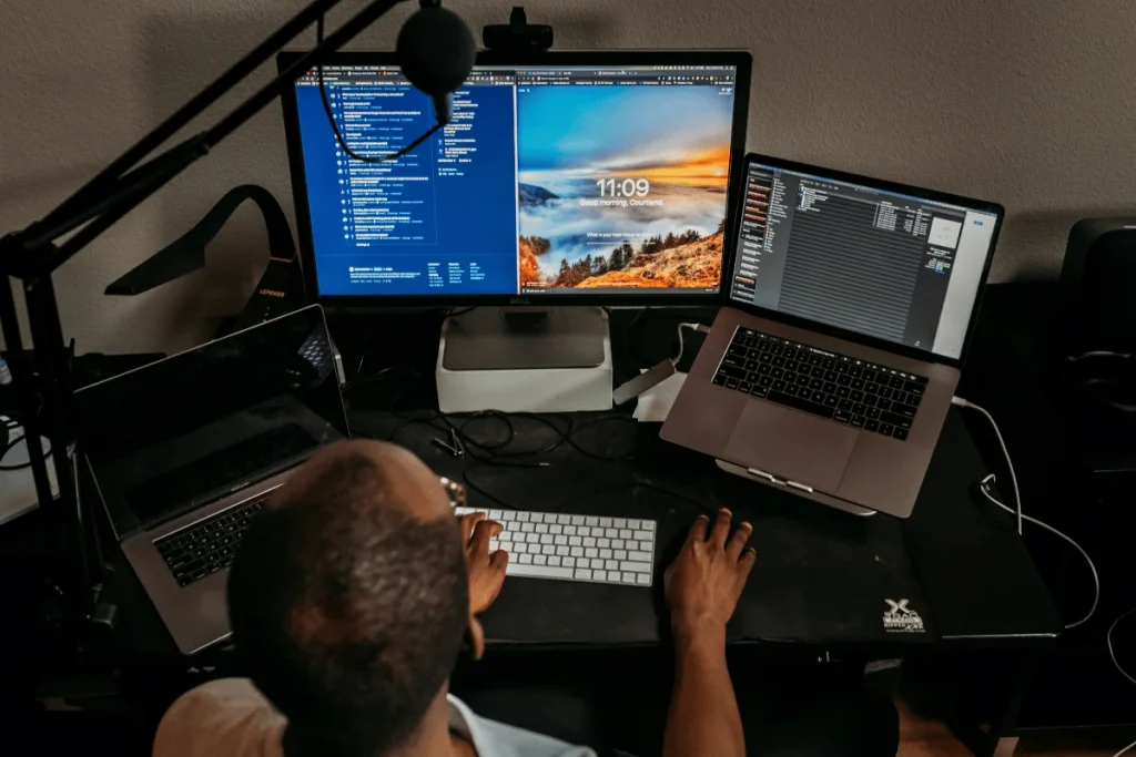 A person works at a desk with two monitors displaying code and a scenic wallpaper, alongside a laptop. The setup suggests focus and productivity.