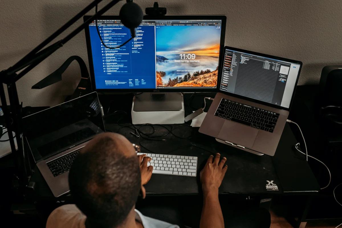A person works at a desk with two monitors displaying code and a scenic wallpaper, alongside a laptop. The setup suggests focus and productivity.