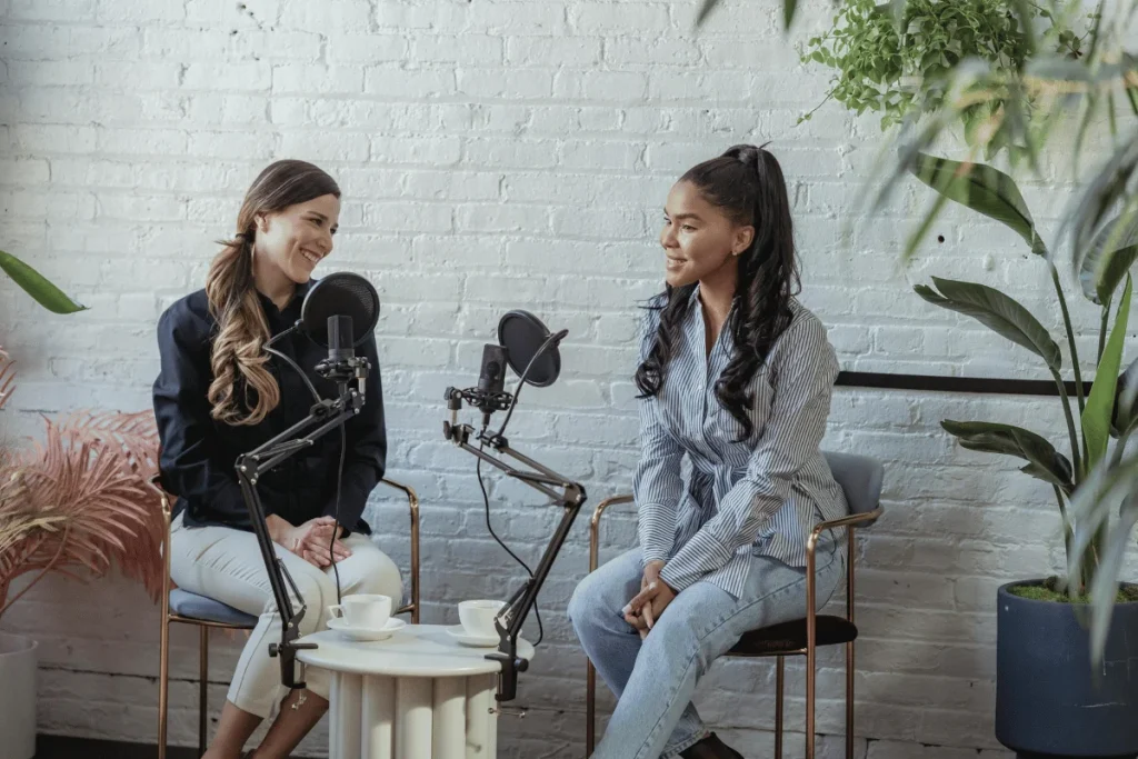 Two women engaged in a podcast recording session with professional microphones in a cozy, plant-filled studio.