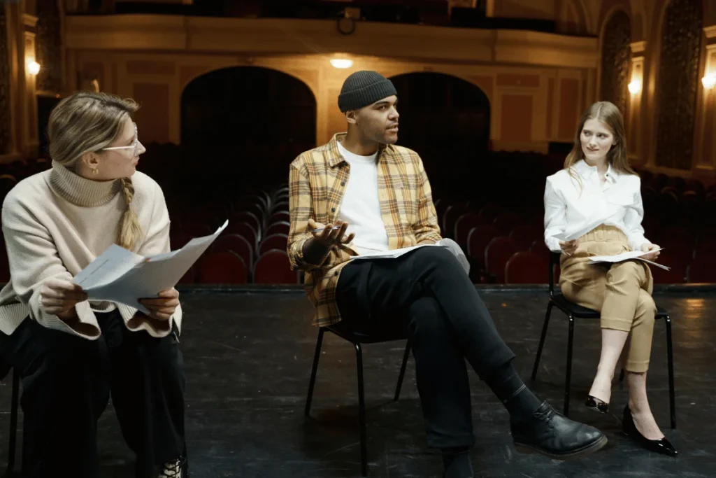 Three actors sitting on stage, holding scripts, and discussing their performance during a theater rehearsal.