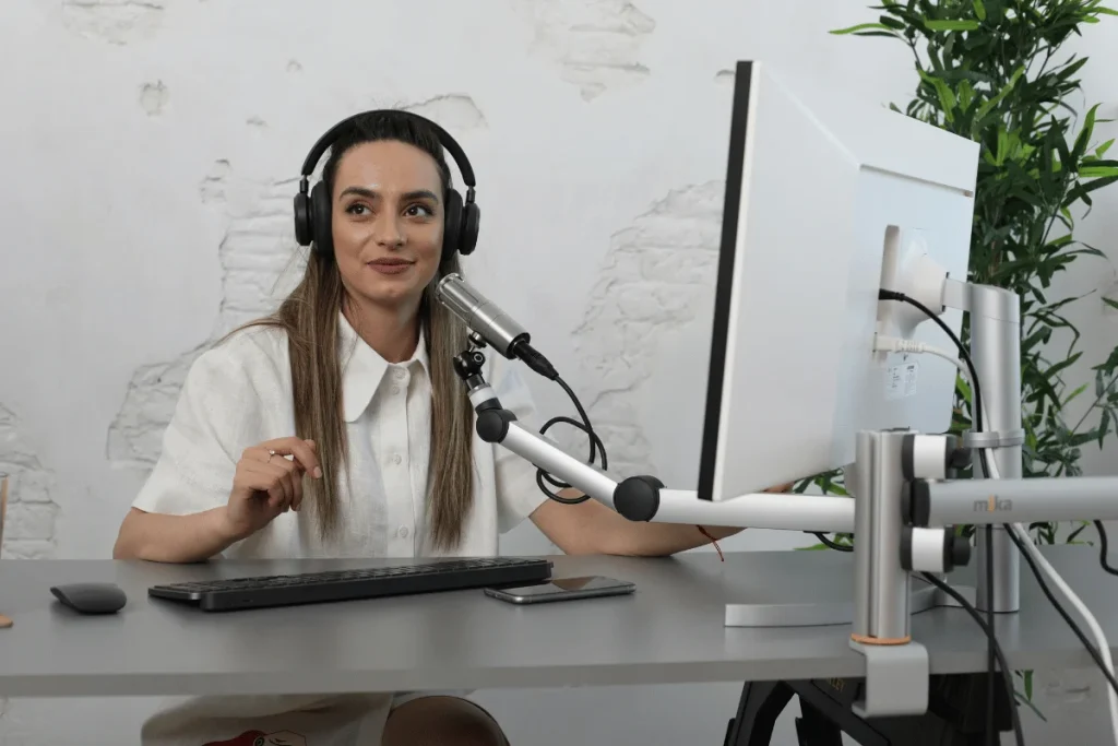 Woman hosting a podcast at a desk with microphone, headphones, and computer monitor in a modern studio.