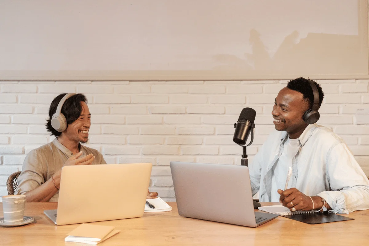 Two men with headphones and laptops hosting a podcast in a cozy studio with a brick wall.