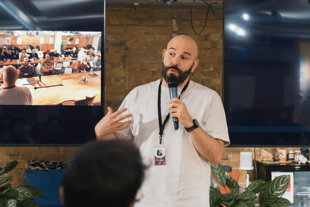 Speaker holding a microphone presenting to an audience in a modern event space with screens displaying a meeting.