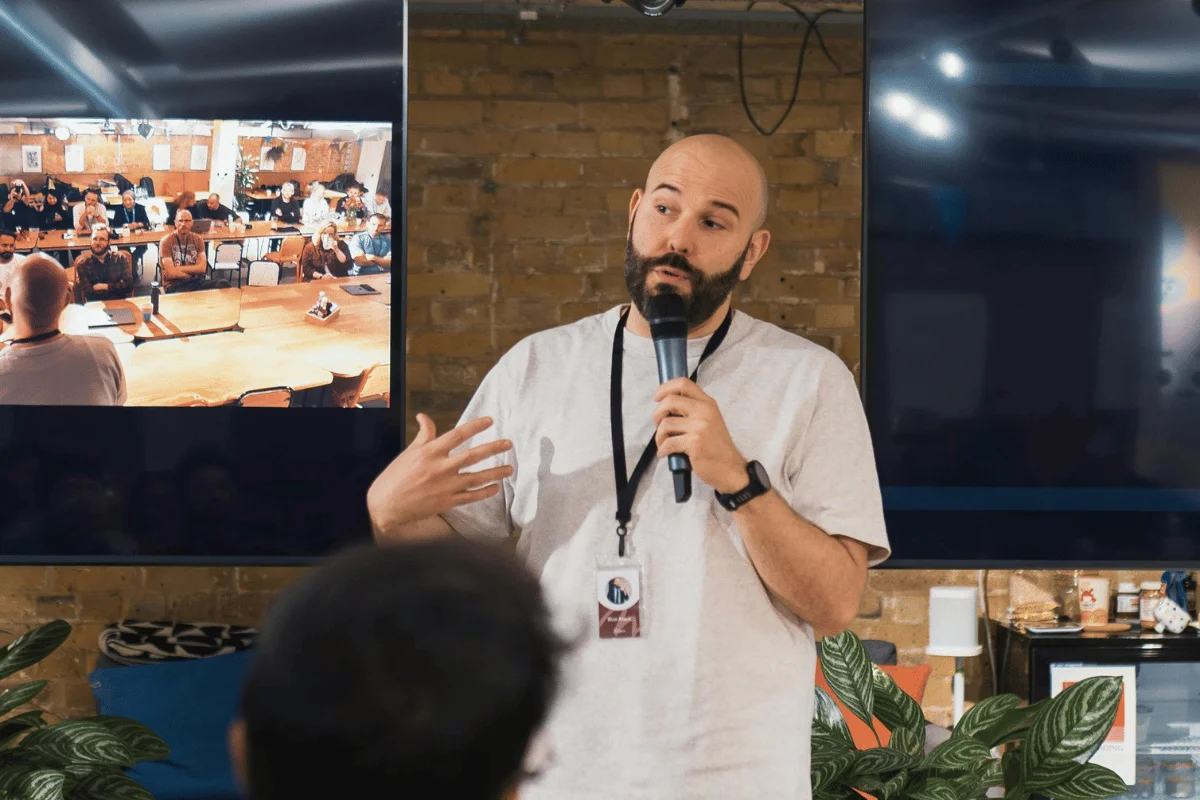 Speaker holding a microphone presenting to an audience in a modern event space with screens displaying a meeting.