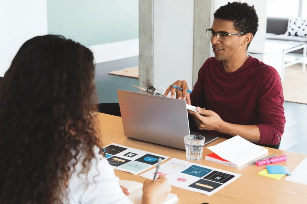 A man and woman collaborate at a table with a laptop, papers, and colorful stationery.