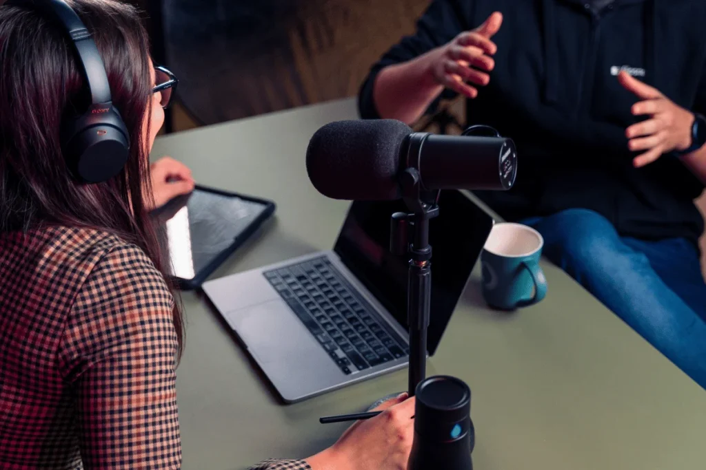 Two podcasters recording at a desk with laptop and mic.