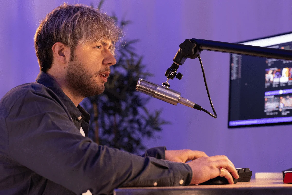 Man recording a podcast with a high-quality microphone and a computer setup in a purple-lit room.