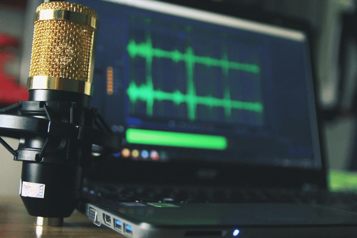 Gold condenser microphone set up in front of a laptop displaying audio waveforms for podcast or voice recording.