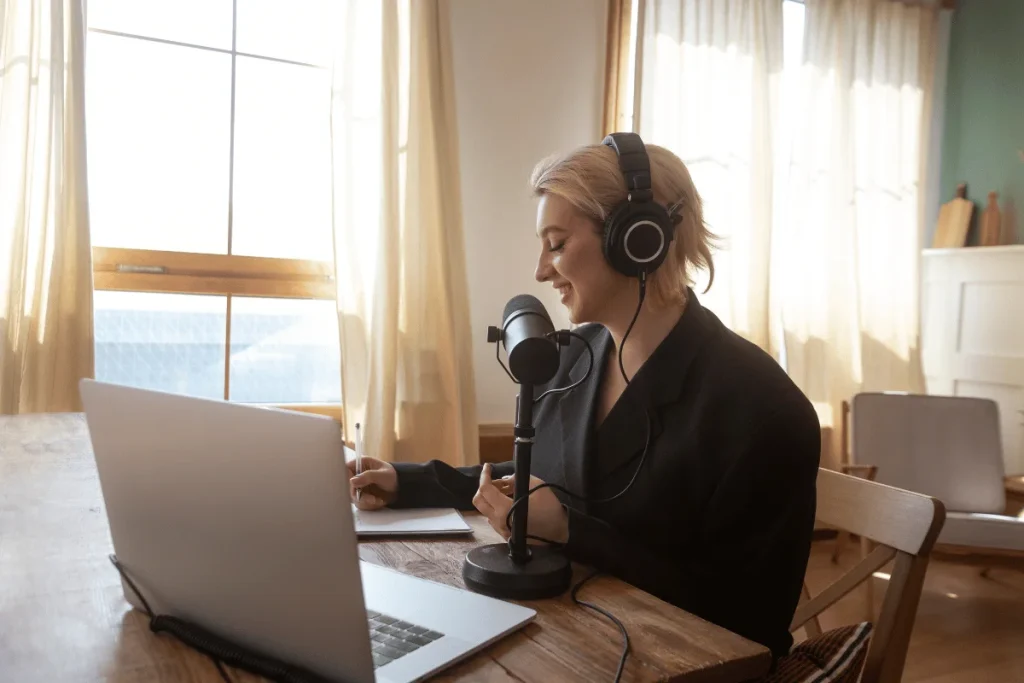 Woman recording a podcast at home using a microphone and headphones while working on a laptop in a sunlit room.