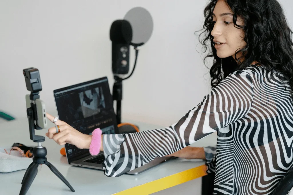 A woman focused on editing a video on her computer while pointing at her camera on a tripod.