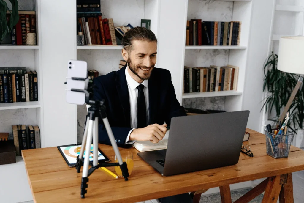 A man dressed in a suit, sitting at a desk with a laptop and tablet, smiling during a virtual meeting.