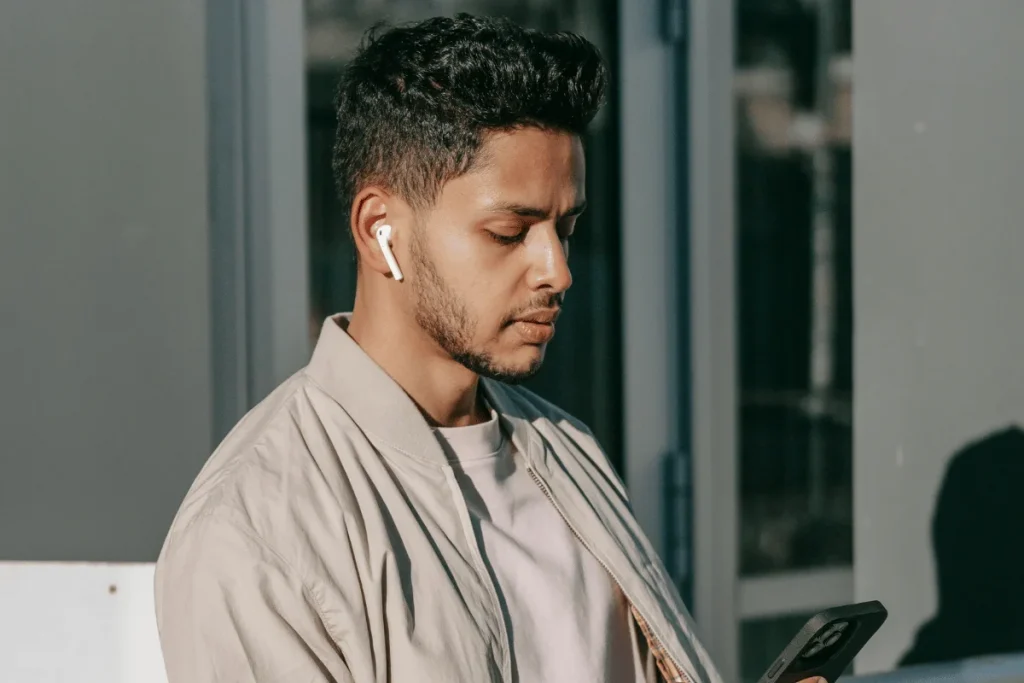 Young man in casual attire with wireless earbuds using a smartphone, urban backdrop.