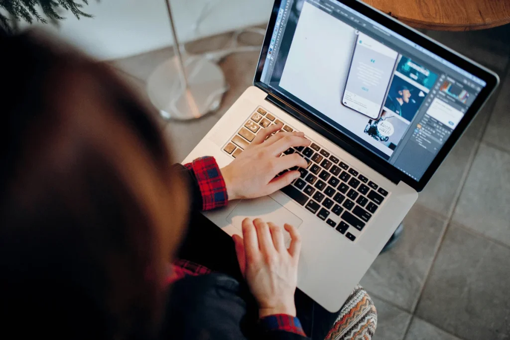 A person in a red plaid shirt uses a laptop, focusing on design software displaying a smartphone mockup.