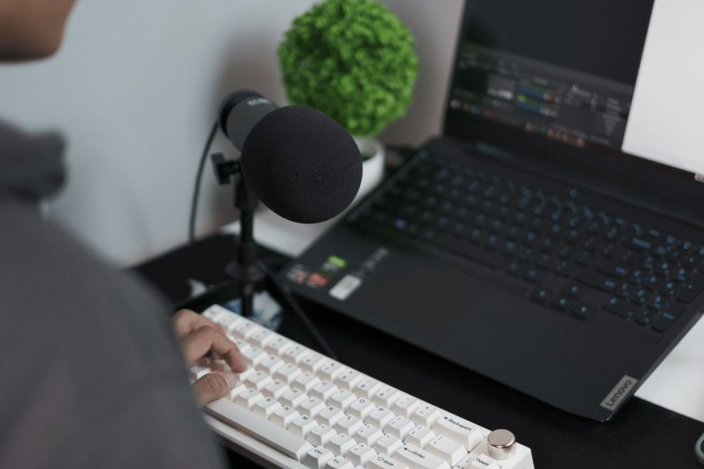 A person types on a white keyboard beside a microphone, with a laptop and a small green plant in the background.
