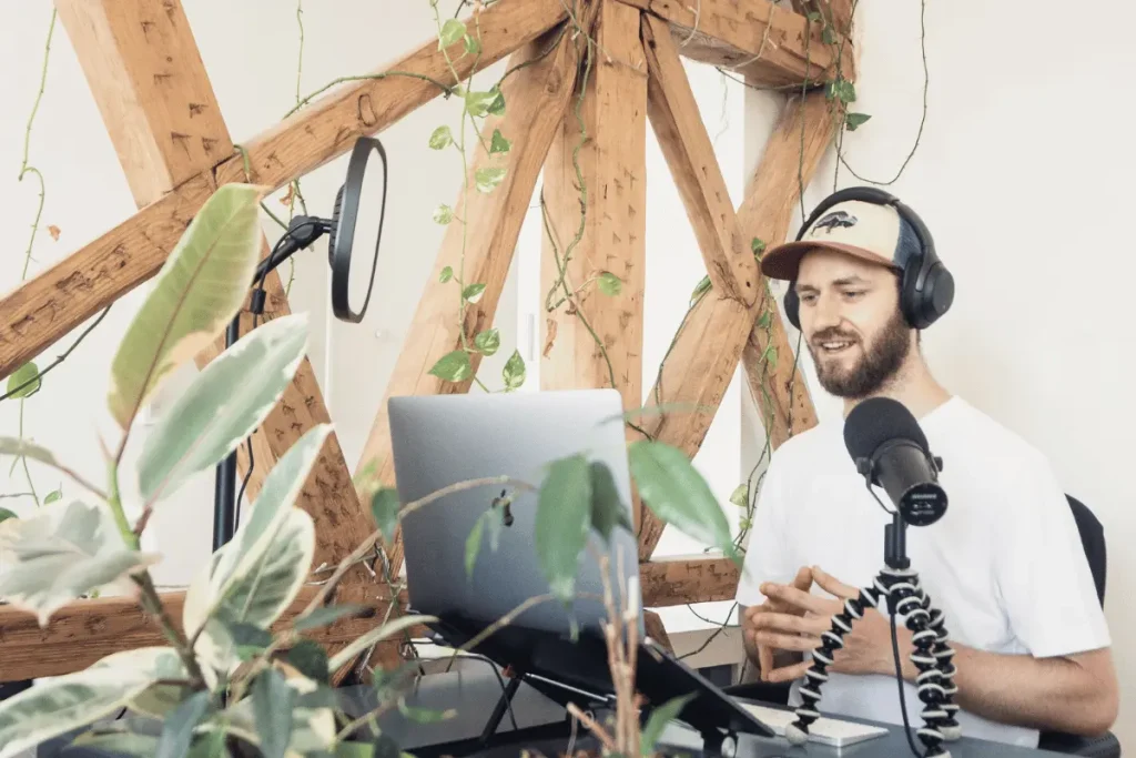 A bearded man in headphones and a cap speaks into a studio microphone beside a laptop, surrounded by leafy plants and rustic wooden beams; the scene feels relaxed and creative.
