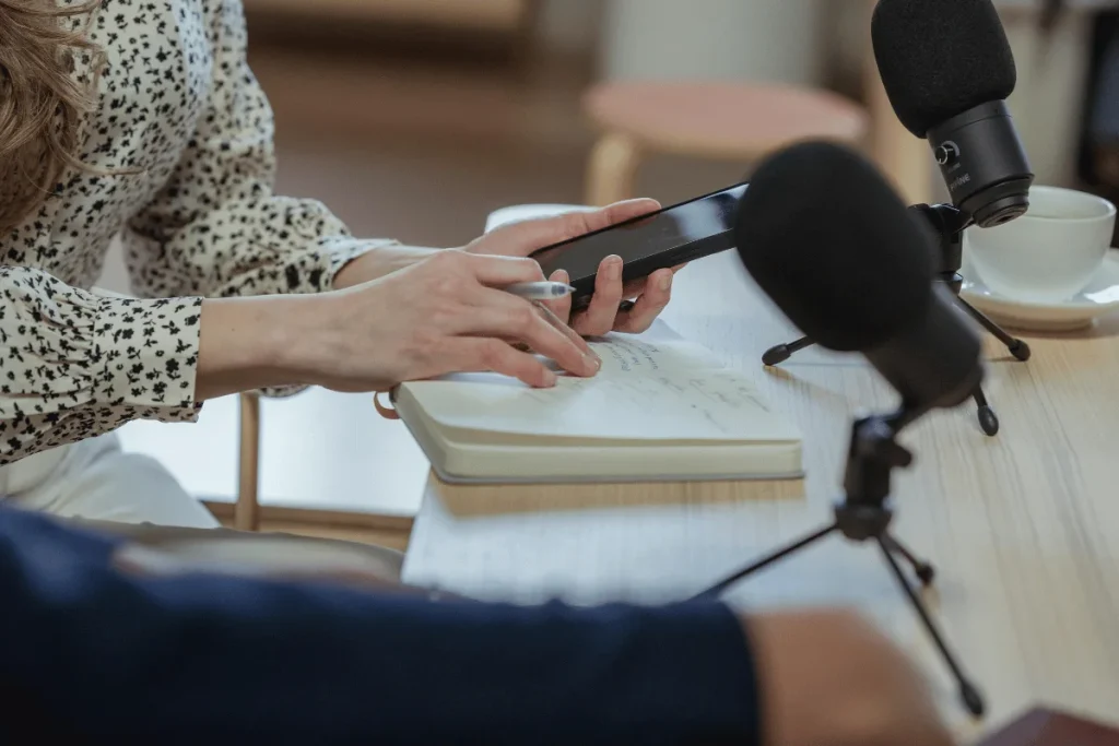 A woman holds a smartphone while taking notes on a notepad during a podcast recording.