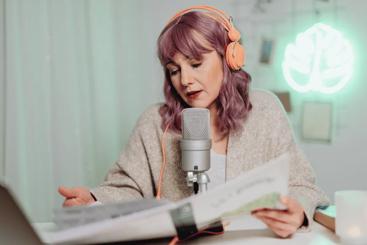 A woman with purple hair and orange headphones reads from a script into a studio microphone, focused in a softly lit recording space.