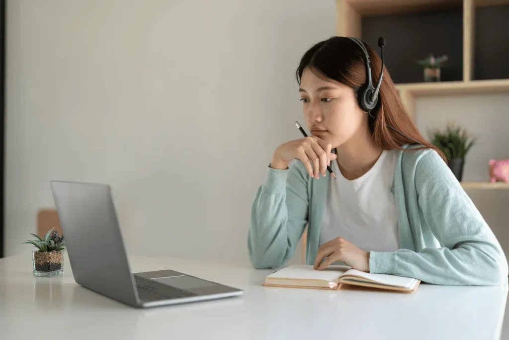 A young woman wearing a headset and holding a pencil while looking thoughtfully at her laptop, possibly engaged in an online meeting.