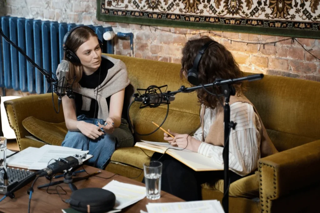 Podcast hosts in casual attire, recording an episode in a relaxed, brick-walled studio environment.