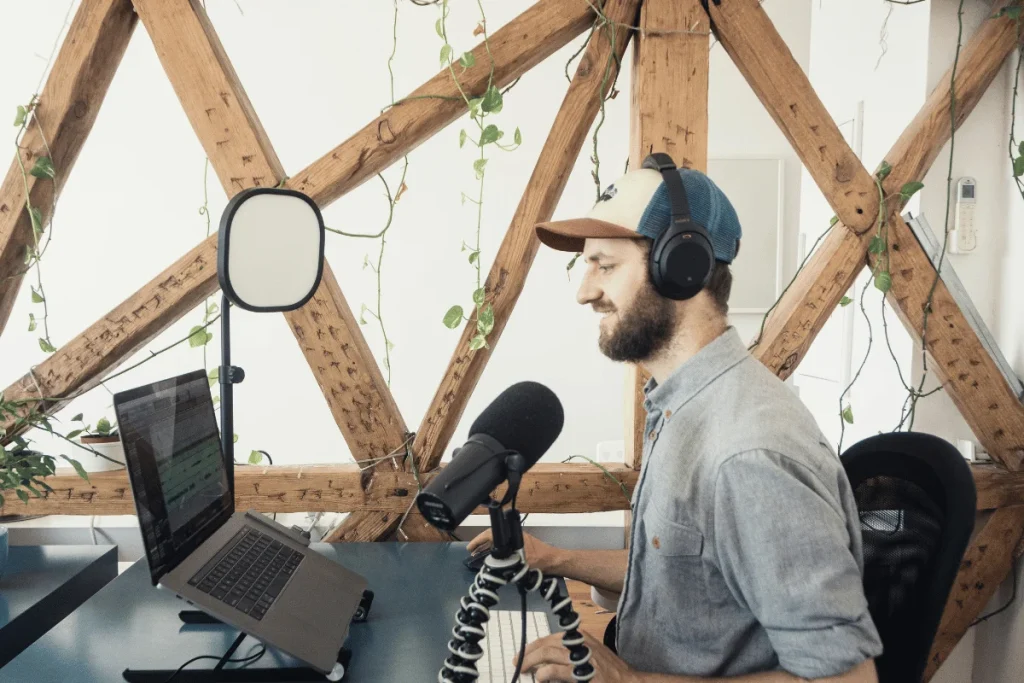 A person wearing headphones speaks into a microphone at a desk with a laptop, backed by a wooden lattice and green vines, conveying a focused atmosphere.
