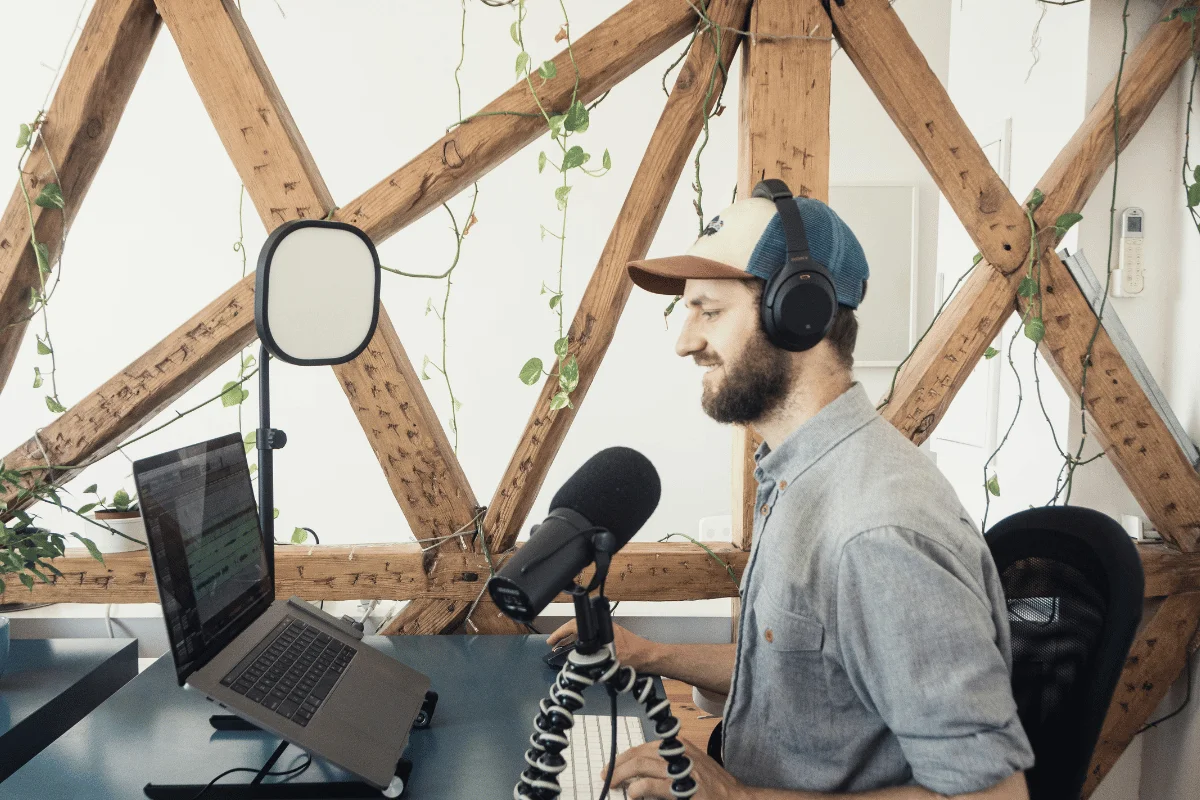 A person wearing headphones speaks into a microphone at a desk with a laptop, backed by a wooden lattice and green vines, conveying a focused atmosphere.