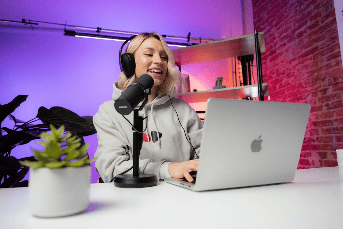 A woman wearing headphones speaks into a microphone while using a laptop in a modern studio.