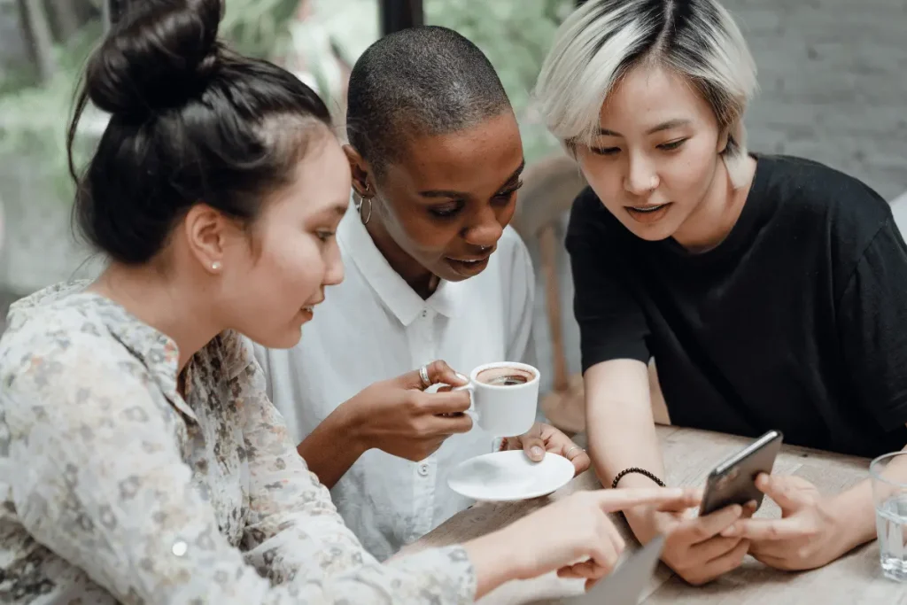 Three friends gather around a smartphone at a café, smiling as they share a moment over coffee. Their relaxed, engaged expressions create a warm, friendly mood.