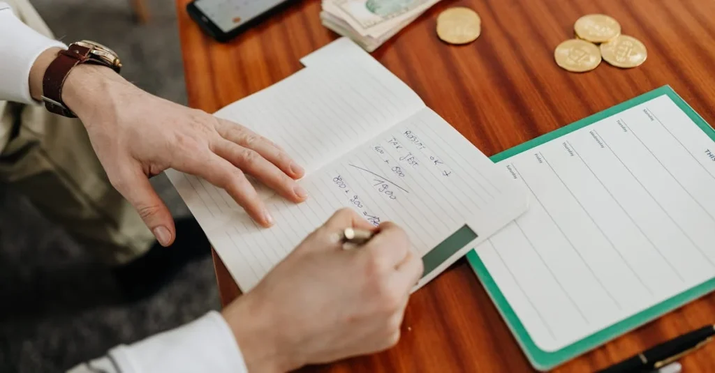 Person writing budget notes on wooden desk with coins, phone, and calculator for podcast production costs.