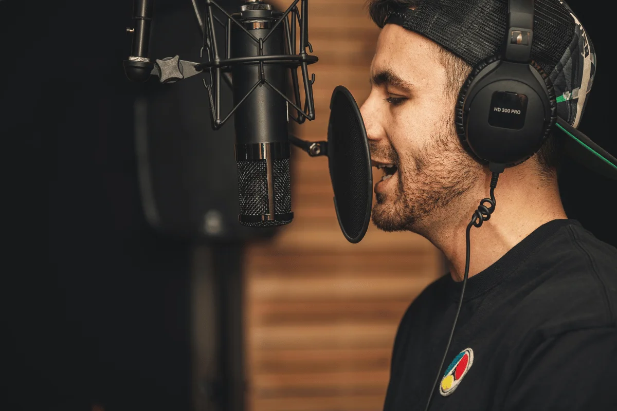 Man wearing headphones and a cap recording vocals into a microphone in a professional studio.
