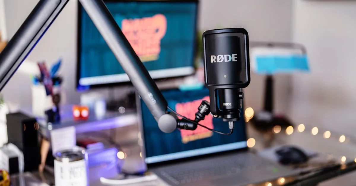RODE microphone mounted on articulating arm in podcast studio with Mac monitor displaying audio waveform editing software.