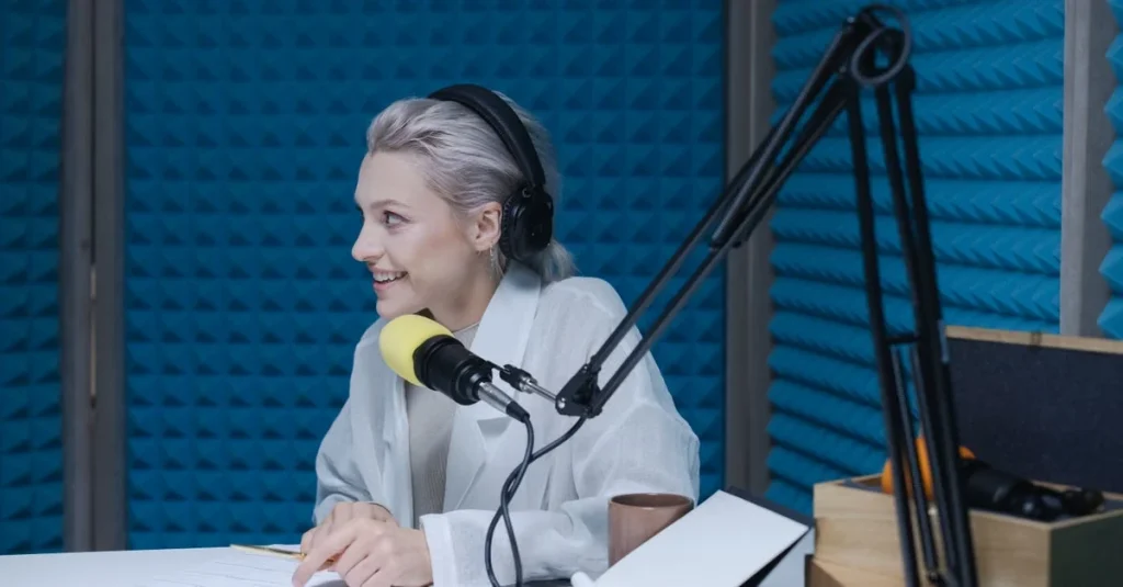 Woman speaking into microphone at podcast hosting studio with blue acoustic panels in background.