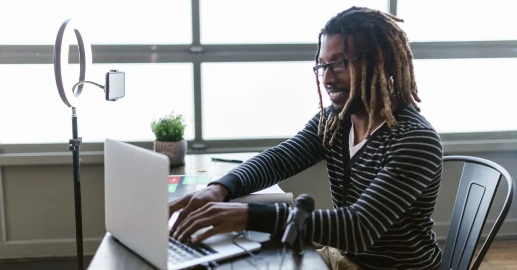 Content creator with dreadlocks at desk with laptop and ring light, comparing Youtube membership vs Patreon monetization.