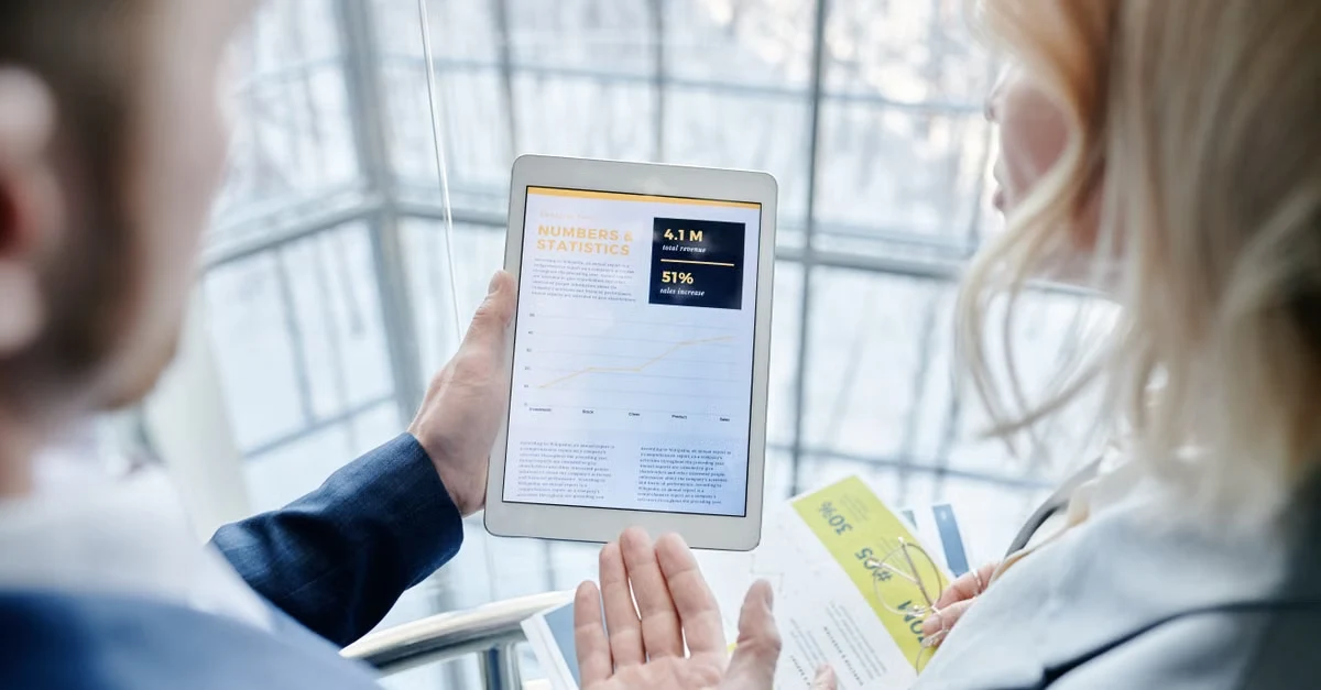 Two people reviewing membership comparison data on tablet with analytics dashboard displayed.