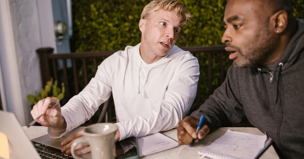 Two men discussing private podcast hosting strategy at desk with laptop and notes, engaged in conversation.