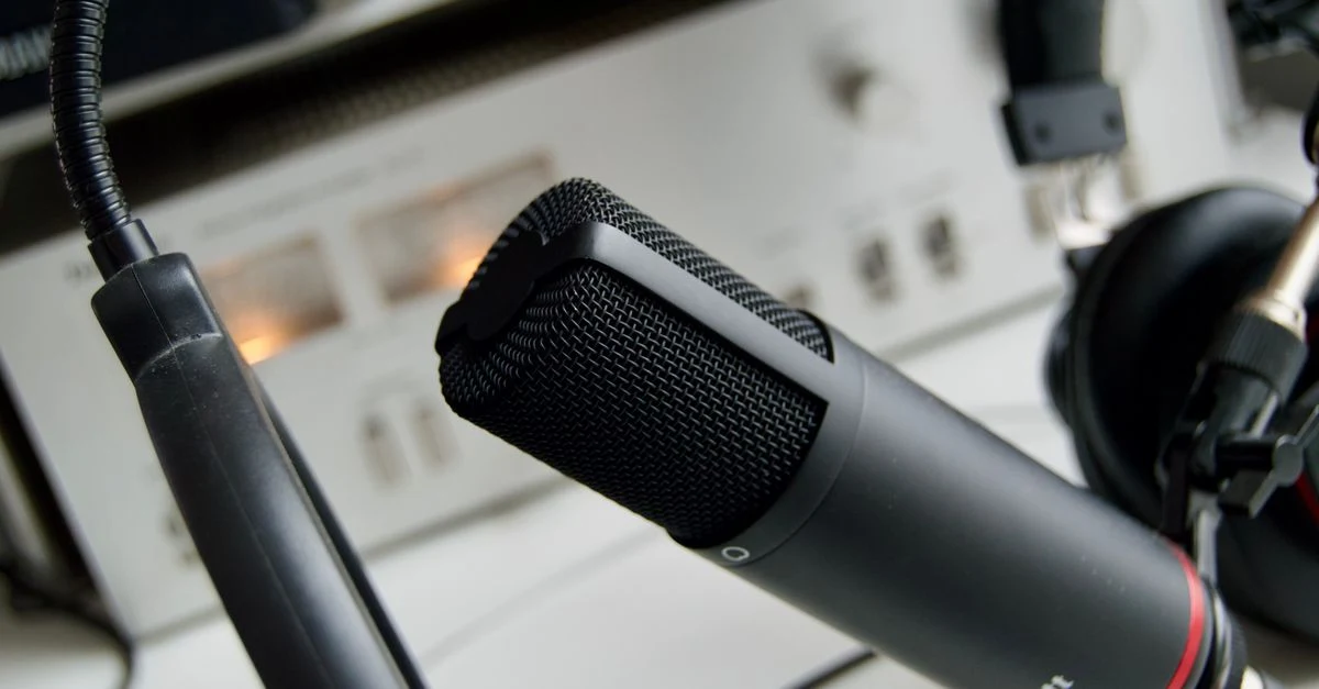 Close-up of a black studio microphone angled across an audio mixing console, with knobs, cables, and controls softly blurred in the background; a focused, professional mood.
