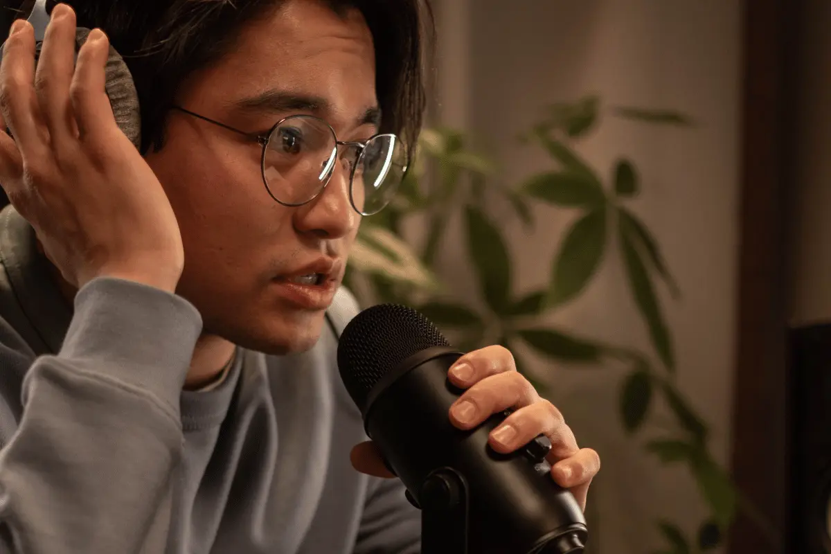 A young man in glasses and a gray sweatshirt cups his ear while speaking into a black microphone, focused in a warmly lit room with plants.