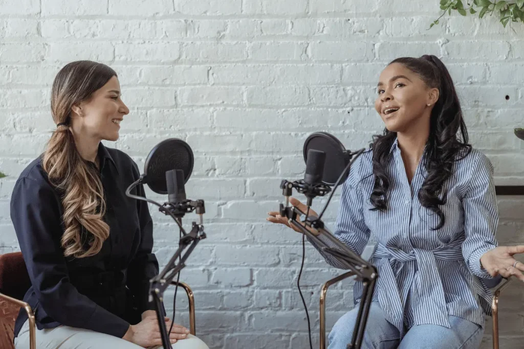 Two women sit facing each other at microphones in a bright podcast studio; one speaks animatedly while the other listens, creating a warm, conversational mood.
