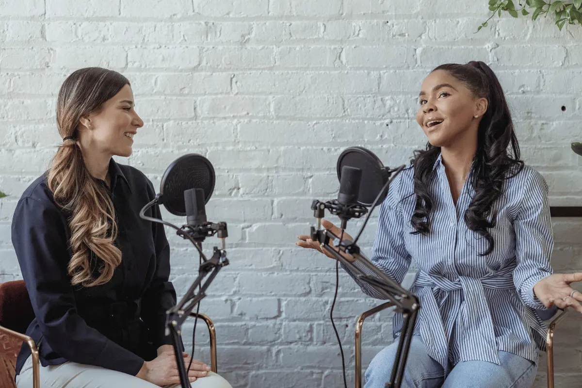 Two women sit facing each other at microphones in a bright podcast studio; one speaks animatedly while the other listens, creating a warm, conversational mood.